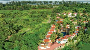 An aerial view of a coastal resort with a linear arrangement of buildings, surrounded by dense tropical vegetation and a clear view of the ocean.