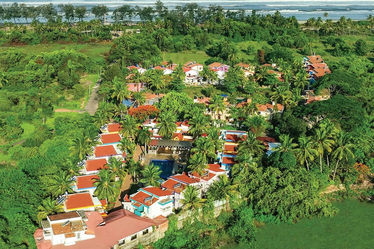 An aerial view of a coastal resort with a linear arrangement of buildings, surrounded by dense tropical vegetation and a clear view of the ocean.