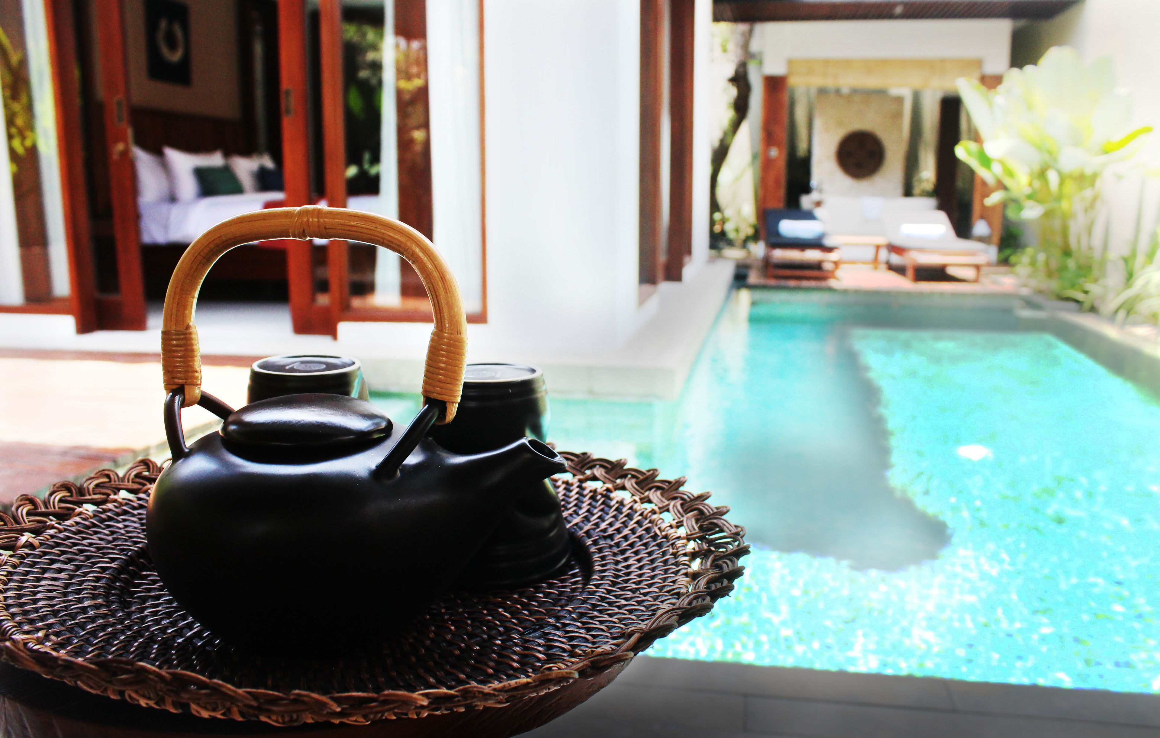A wicker tray with two black teapots and a cup, placed on a balcony overlooking a swimming pool.