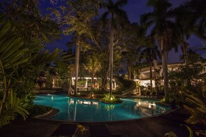 Un área de piscina nocturna con palmeras, sillas de descanso y un edificio al fondo.