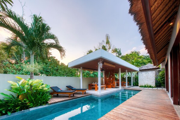 Piscina con terraza de madera, sillas de descanso y zona de estar cubierta.