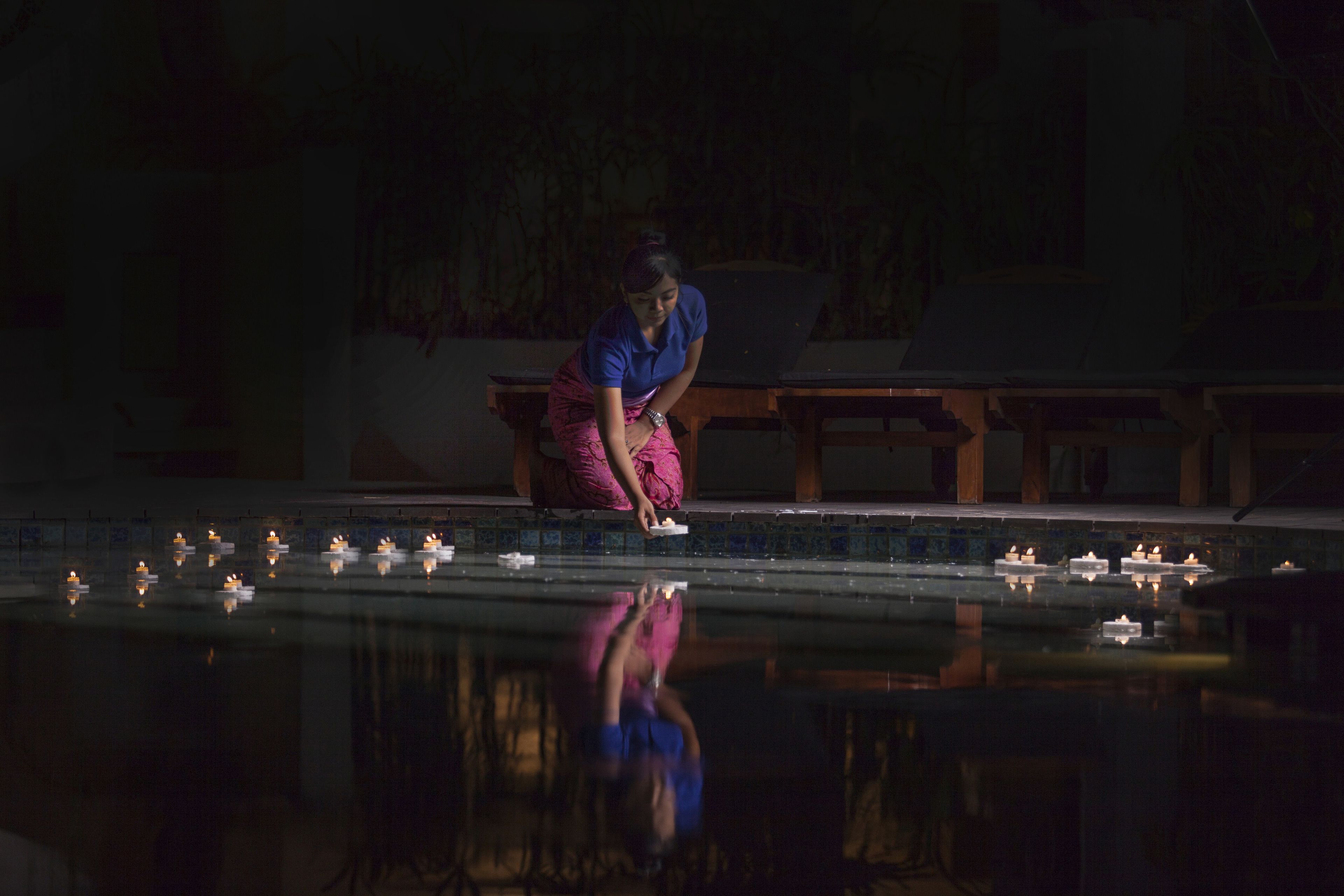 A person in a blue shirt and pink skirt kneeling by a reflective pool with candles.
