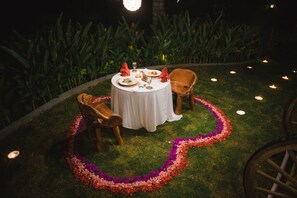 Mesa redonda dispuesta para dos personas, con mantel blanco, bordeada de flores, y otras mesas iluminadas con velas al fondo.