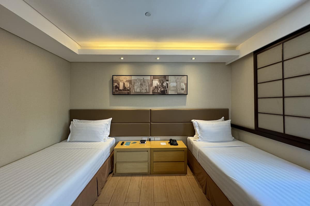 A hotel room with two single beds, a small desk with a chair, a nightstand, and a wall-mounted artwork.