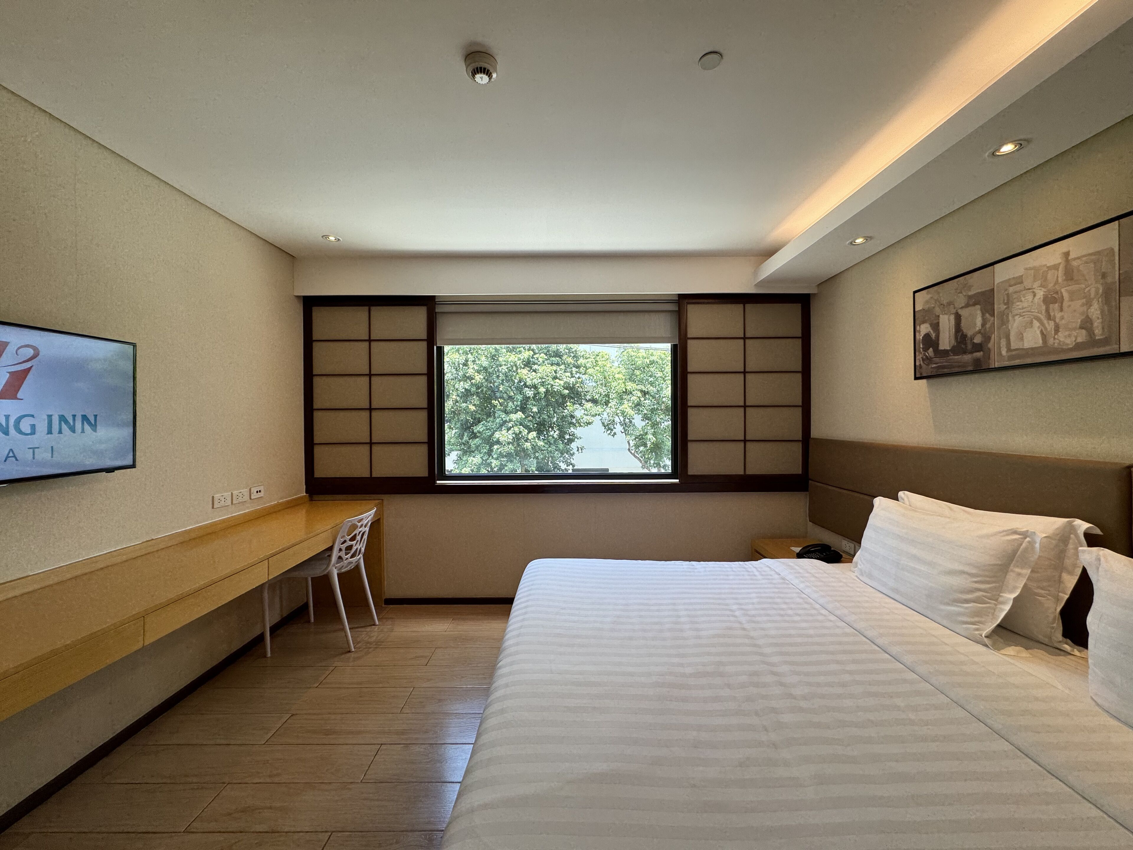 A hotel room with a bed, a desk, a chair, a window with a view of trees, and a wall-mounted TV.