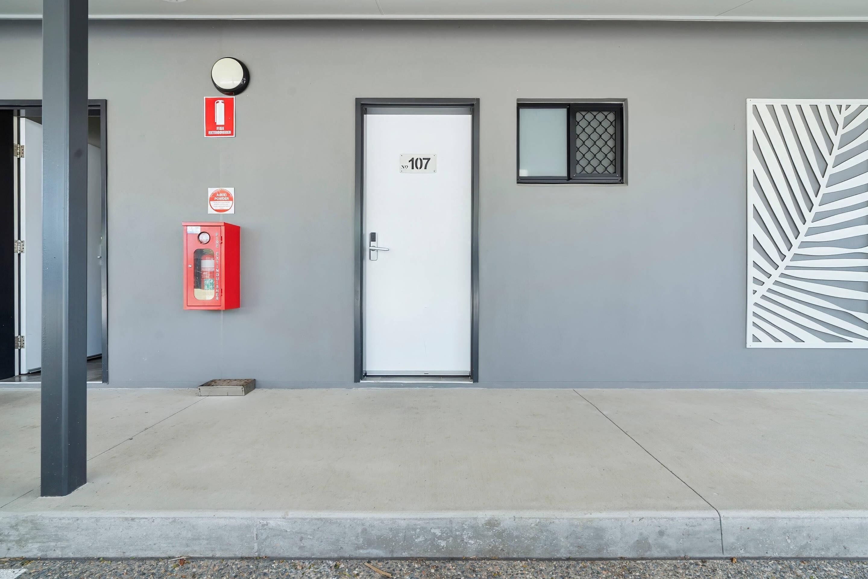 Fachada de edificio con una puerta etiquetada '107', un detector de incendios, una caja roja de emergencia y un panel decorativo.