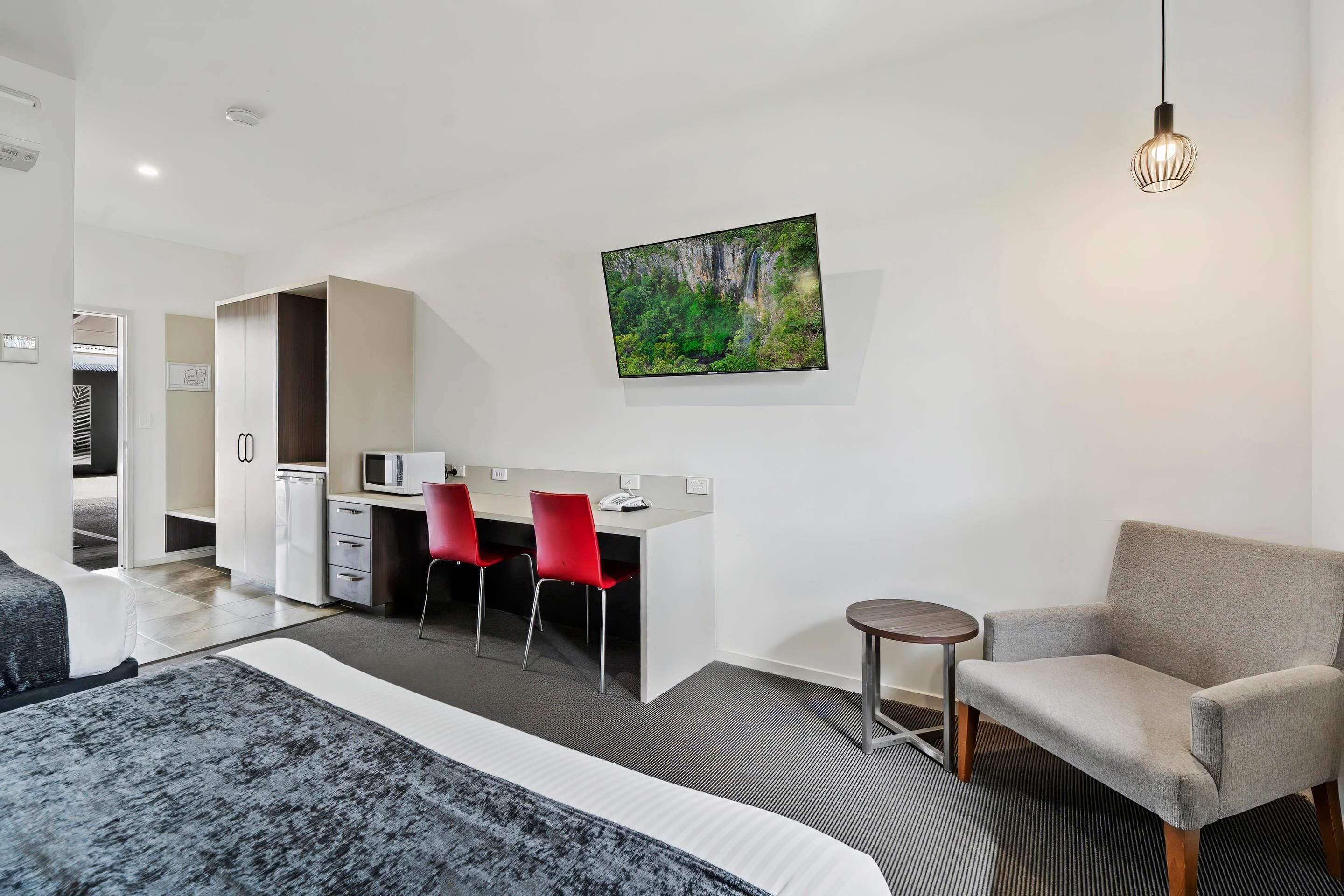 Una habitación de hotel moderna con una cama, un escritorio con dos sillas rojas, una mesita redonda y un cuadro de una cascada en la pared.