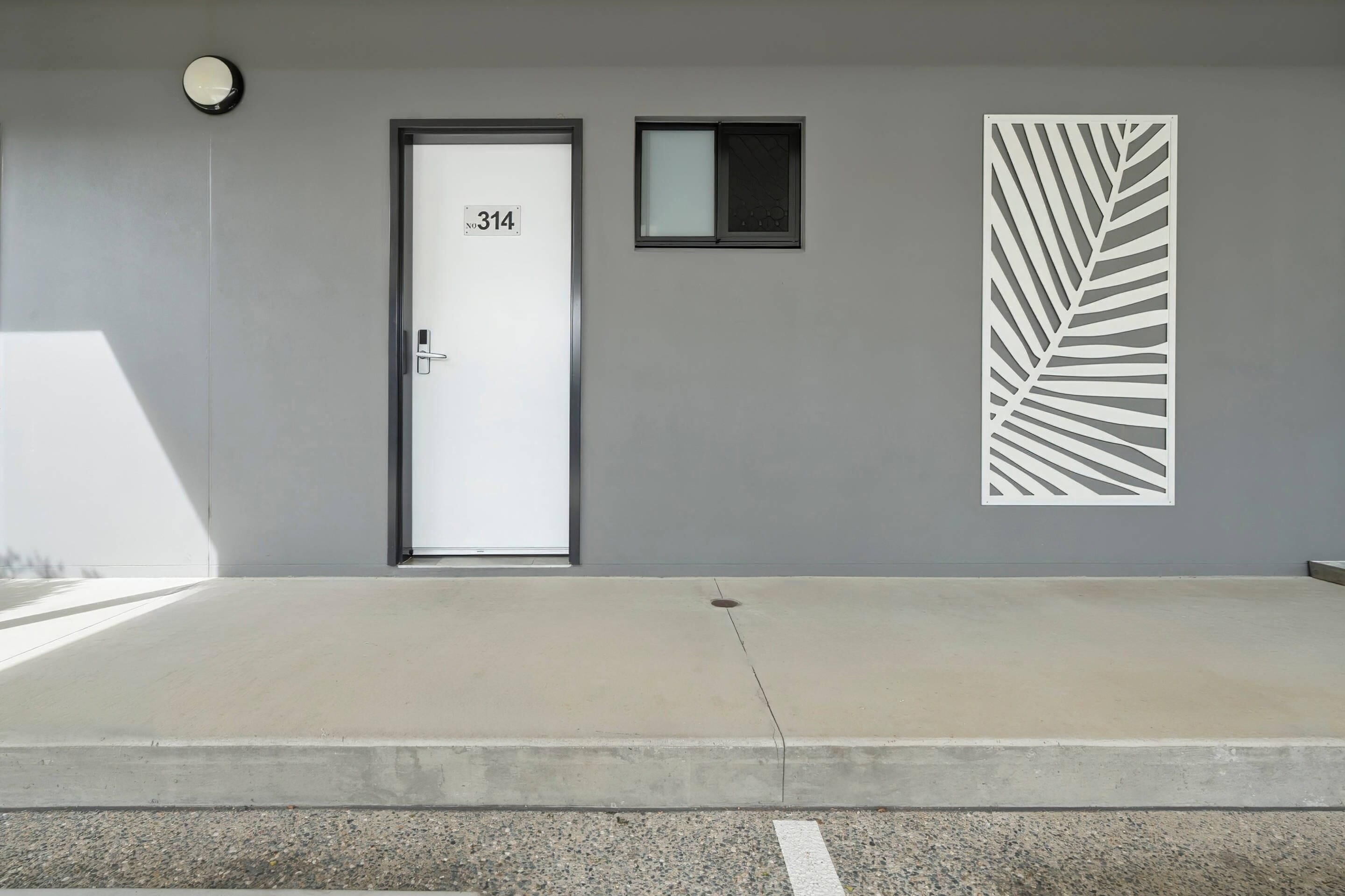 Entrada de un edificio con una puerta identificada con el número '314', un ventanal pequeño encima y un panel decorativo con diseño de hojas en la pared.