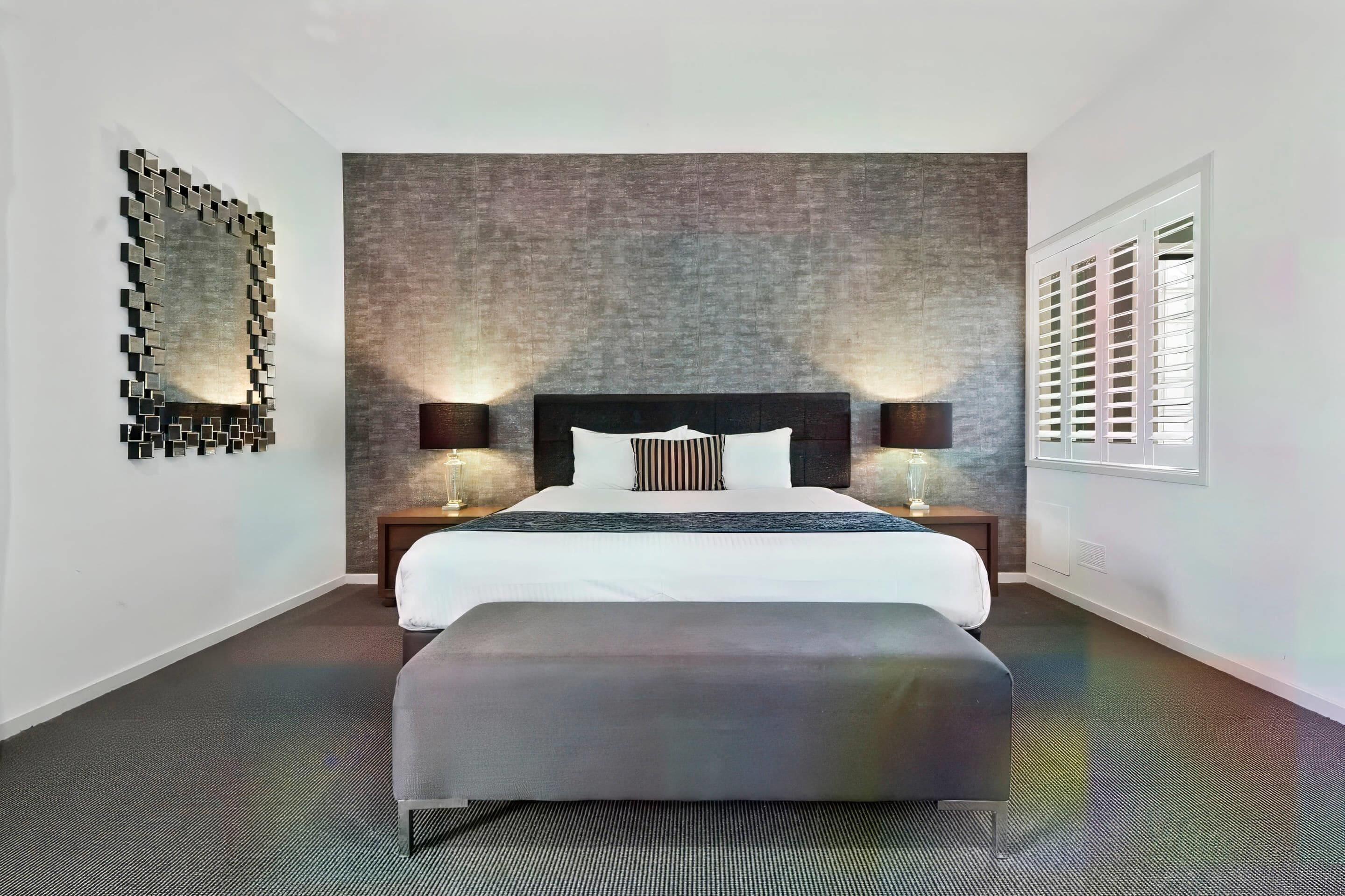 A modern hotel room with a large bed, two bedside tables, a bench, a textured wall, a mirror, and a window with blinds.