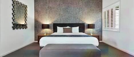 A modern hotel room with a large bed, two bedside tables, a bench, a textured wall, a mirror, and a window with blinds.