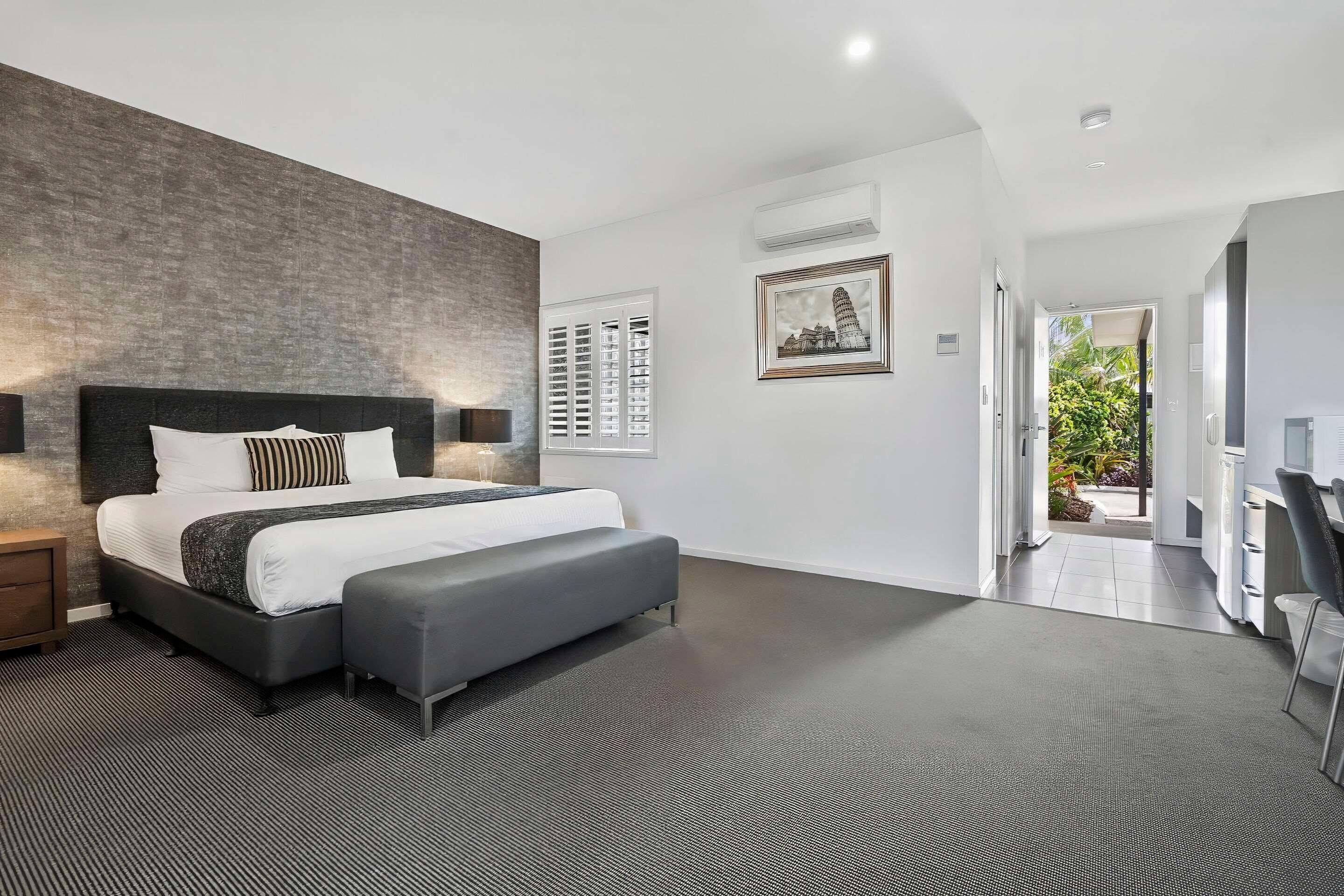 Une chambre moderne avec un grand lit, un banc gris, un mur décoratif en briques, un tableau encadré et une vue sur l’extérieur par une porte ouverte.