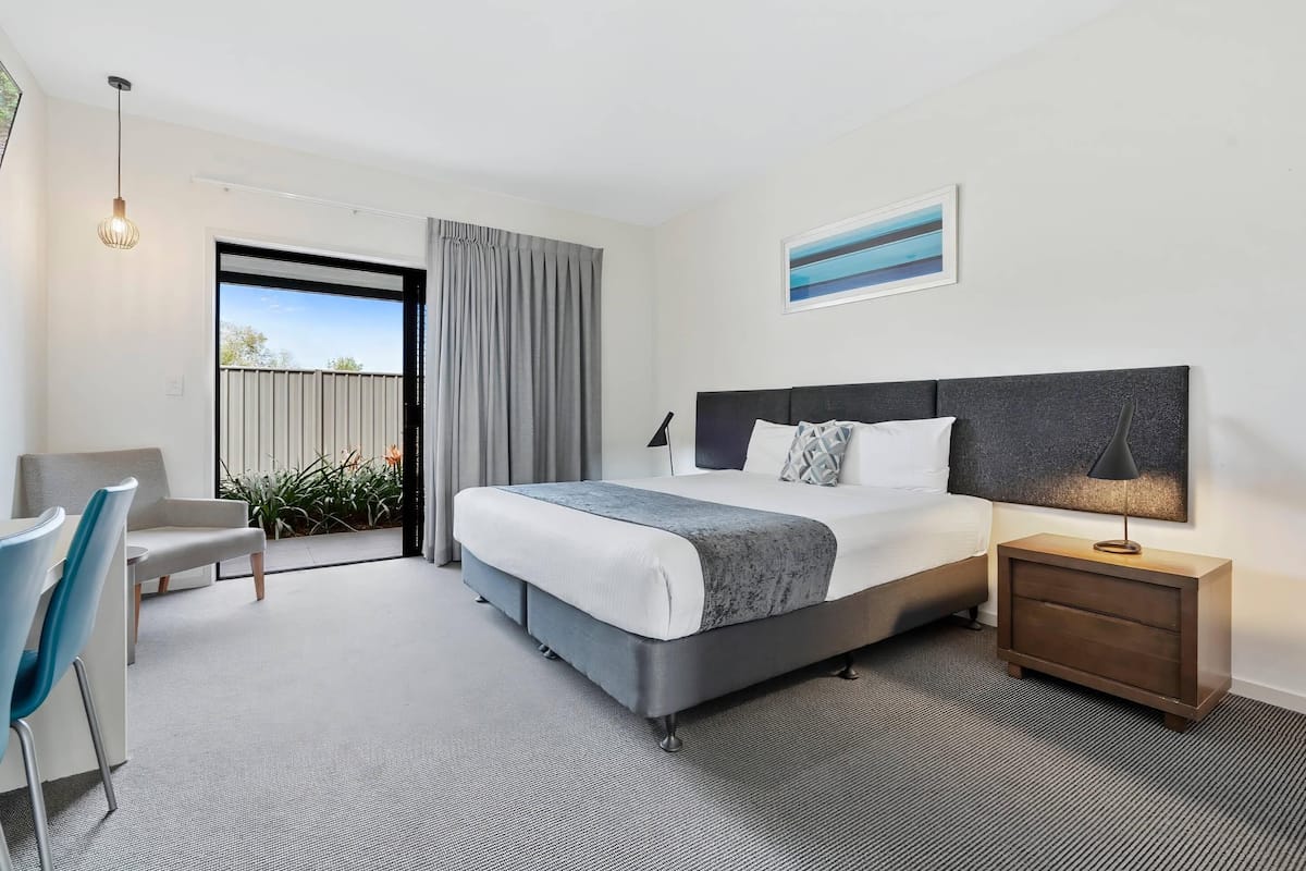 Una habitación de hotel moderna con una cama grande, una mesita de noche con lámpara, una silla y vistas al exterior.