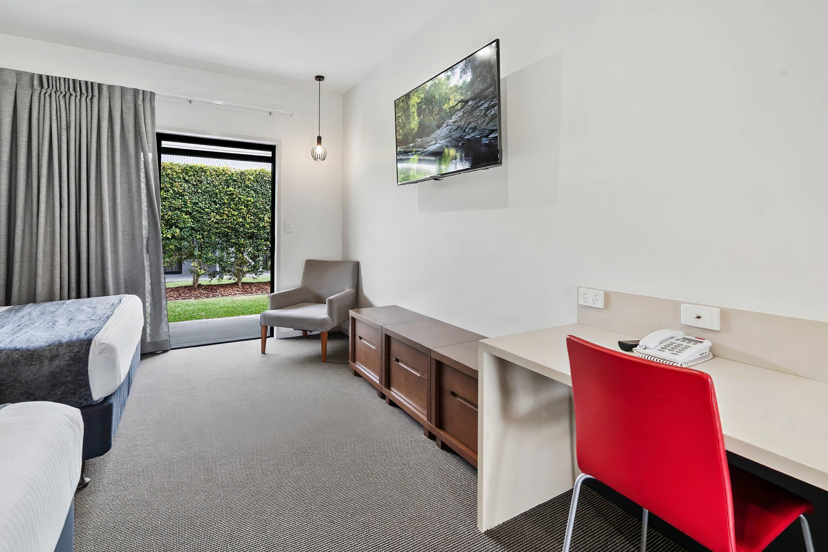A hotel room with a bed, a desk with a telephone, a chair, a TV mounted on the wall, and a view of greenery outside.