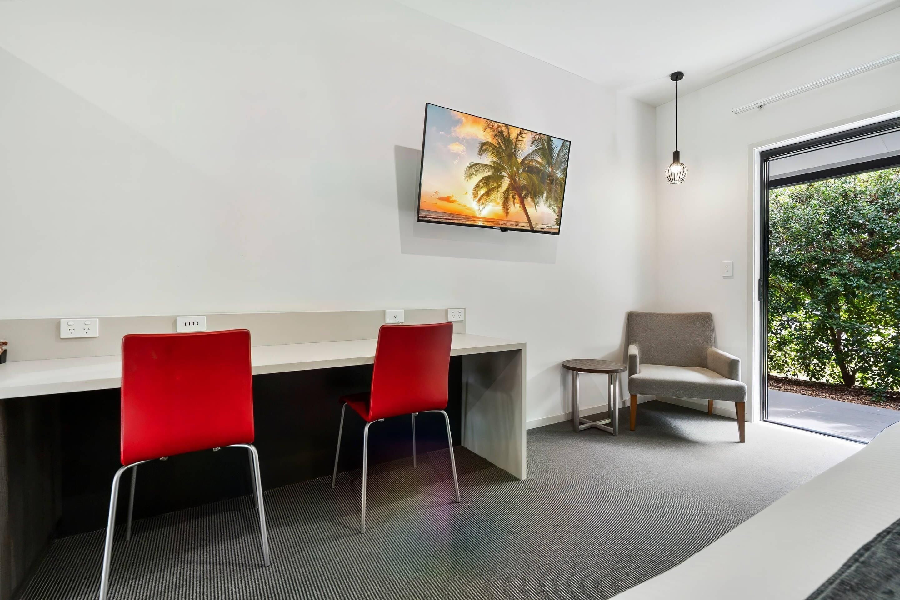 A modern room with a flat-screen TV, a desk with two red chairs, a grey armchair, and a large window with a view of greenery.