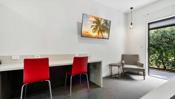A modern room with a flat-screen TV, a desk with two red chairs, a grey armchair, and a large window with a view of greenery.