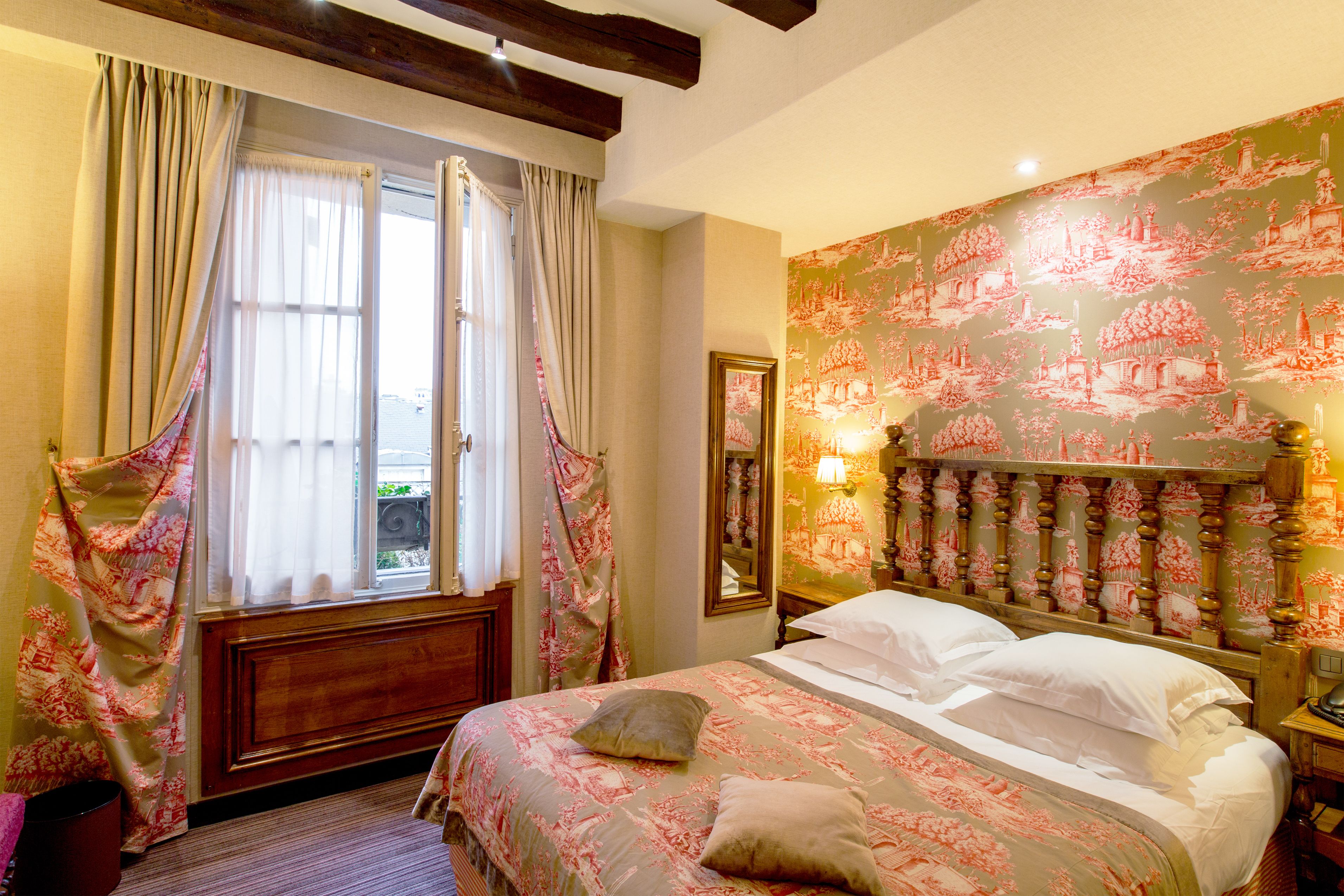 Une chambre avec un grand lit, une fenêtre avec des rideaux, un mur recouvert d’un papier peint à motifs et une table de chevet avec une lampe.