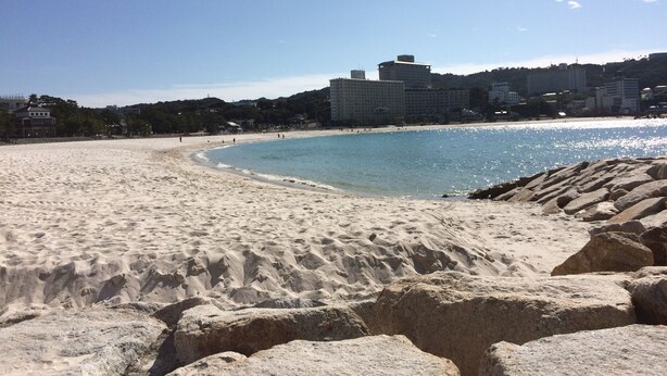 Ein sandiger Strand mit klarem, blauem Wasser, große Felsen im Vordergrund und Gebäude im Hintergrund.