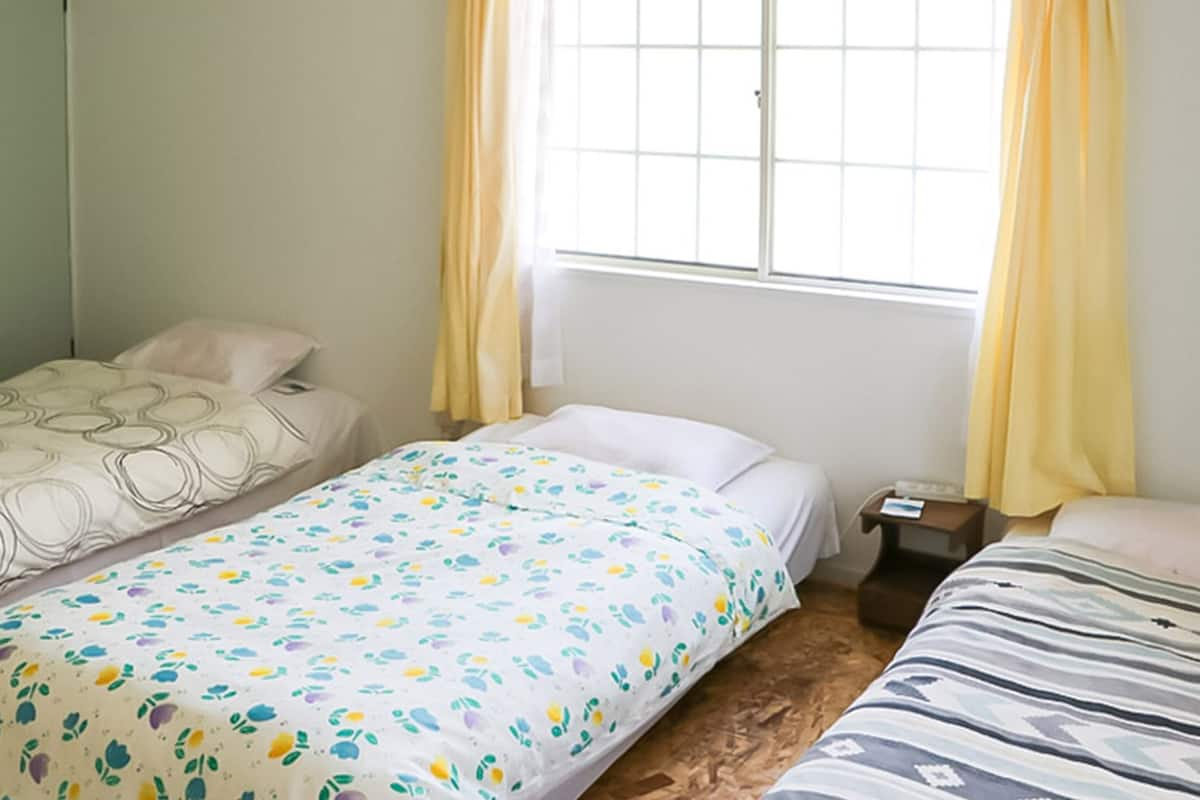 A room with two single beds, a wardrobe, a window with curtains, and a bedside table with a lamp.