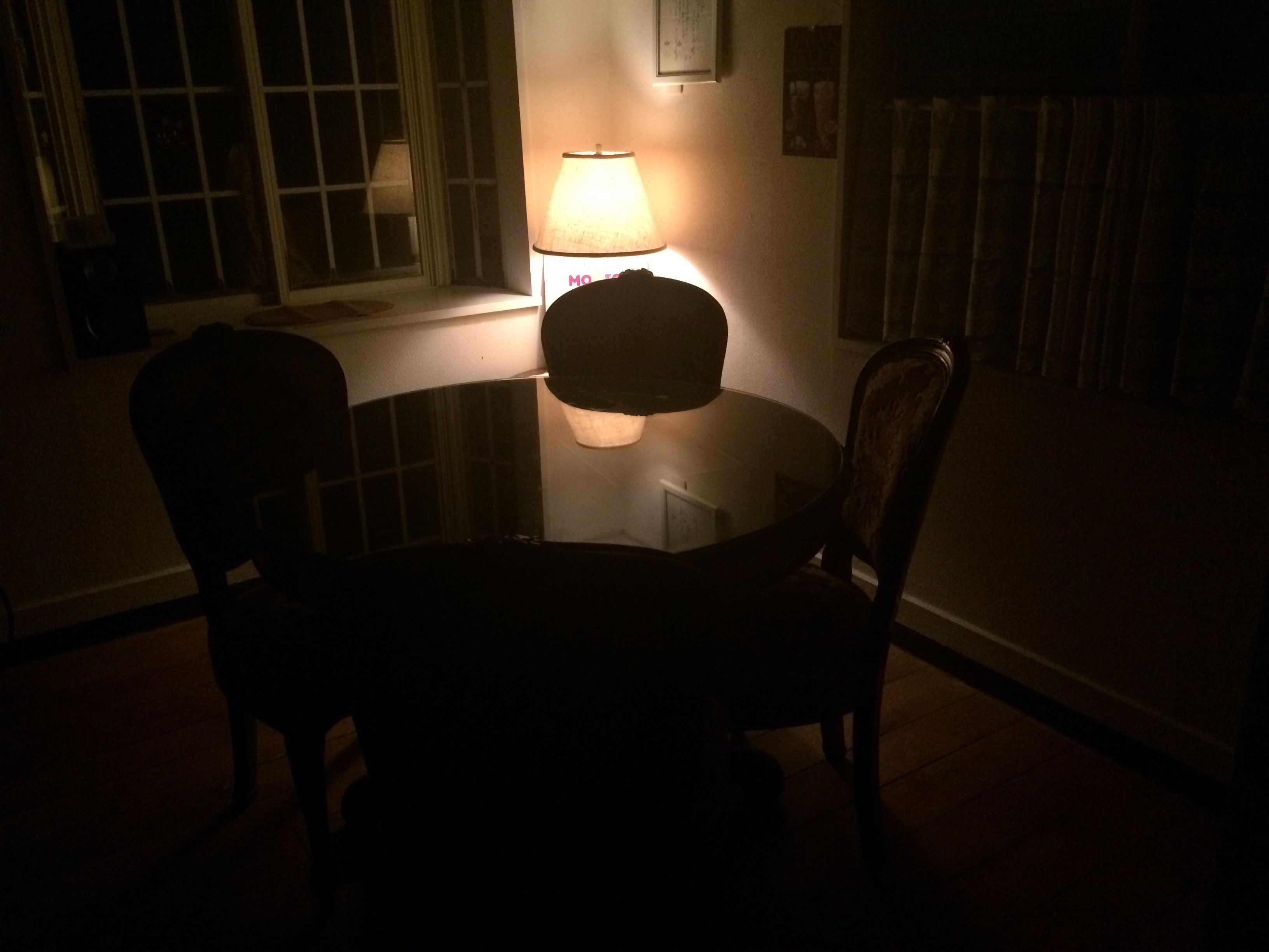 A dimly lit room with a round table and two chairs, a lamp on the table, and a window with blinds.