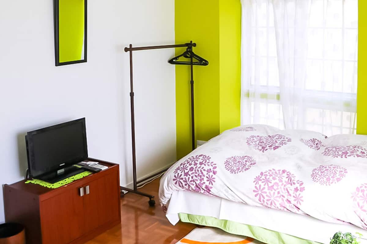 A bedroom with a bed, a TV on a wooden cabinet, a floor lamp, and a window with sheer curtains.