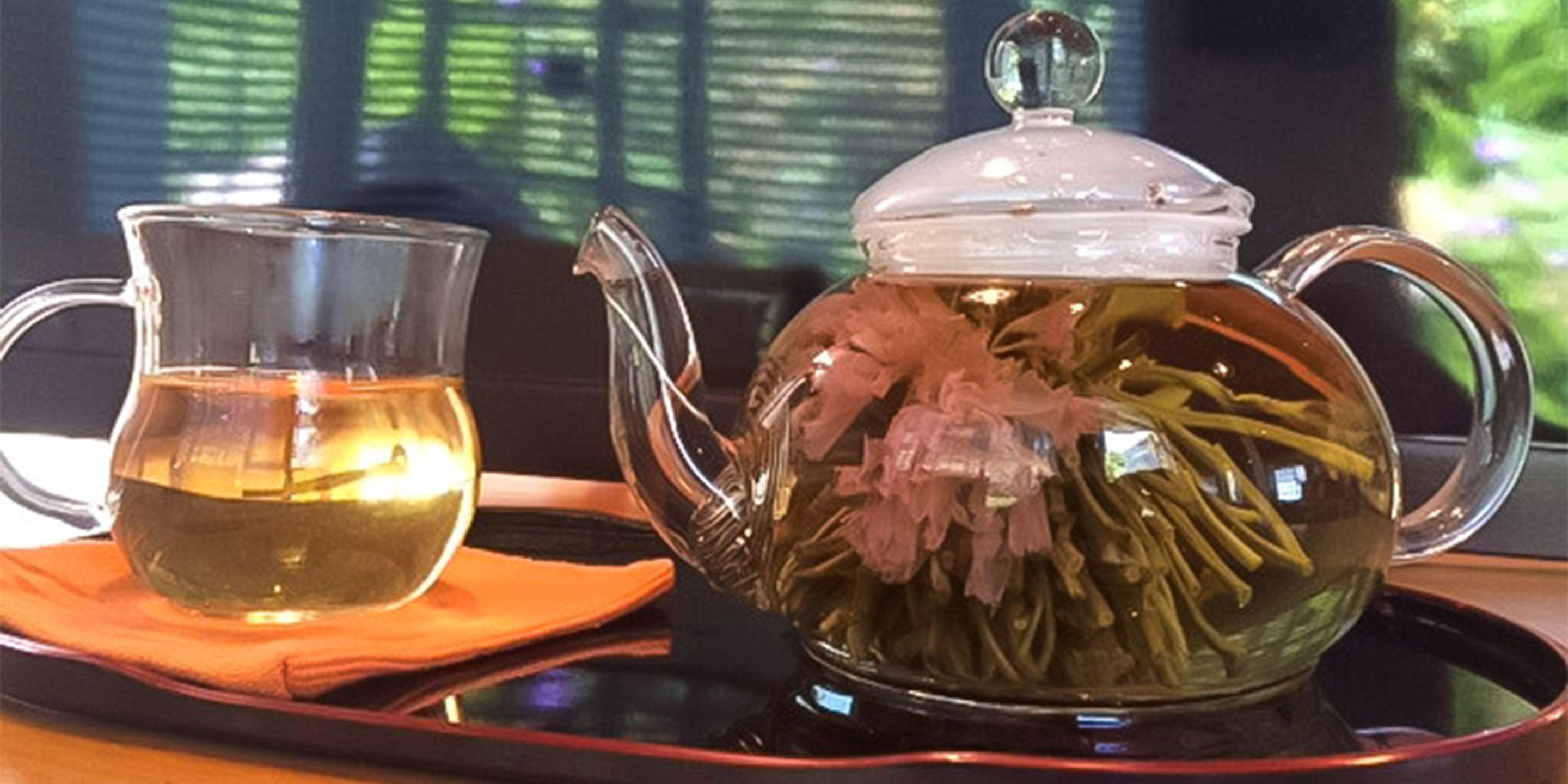 A clear glass teapot with a lid, filled with tea and dried flowers, placed on a tray.