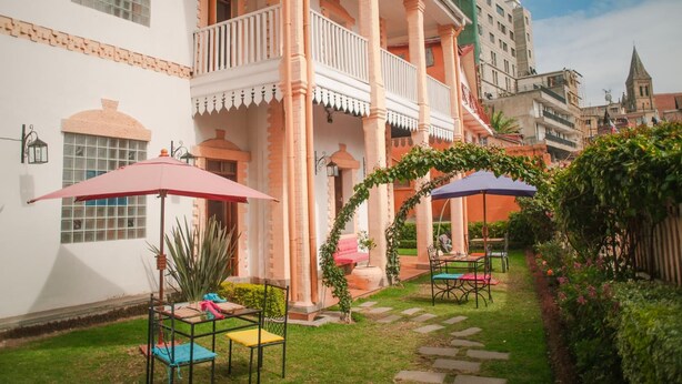 A courtyard with a white building, columns, and a balcony. There are outdoor tables and chairs, and a small garden area with plants and a pathway.