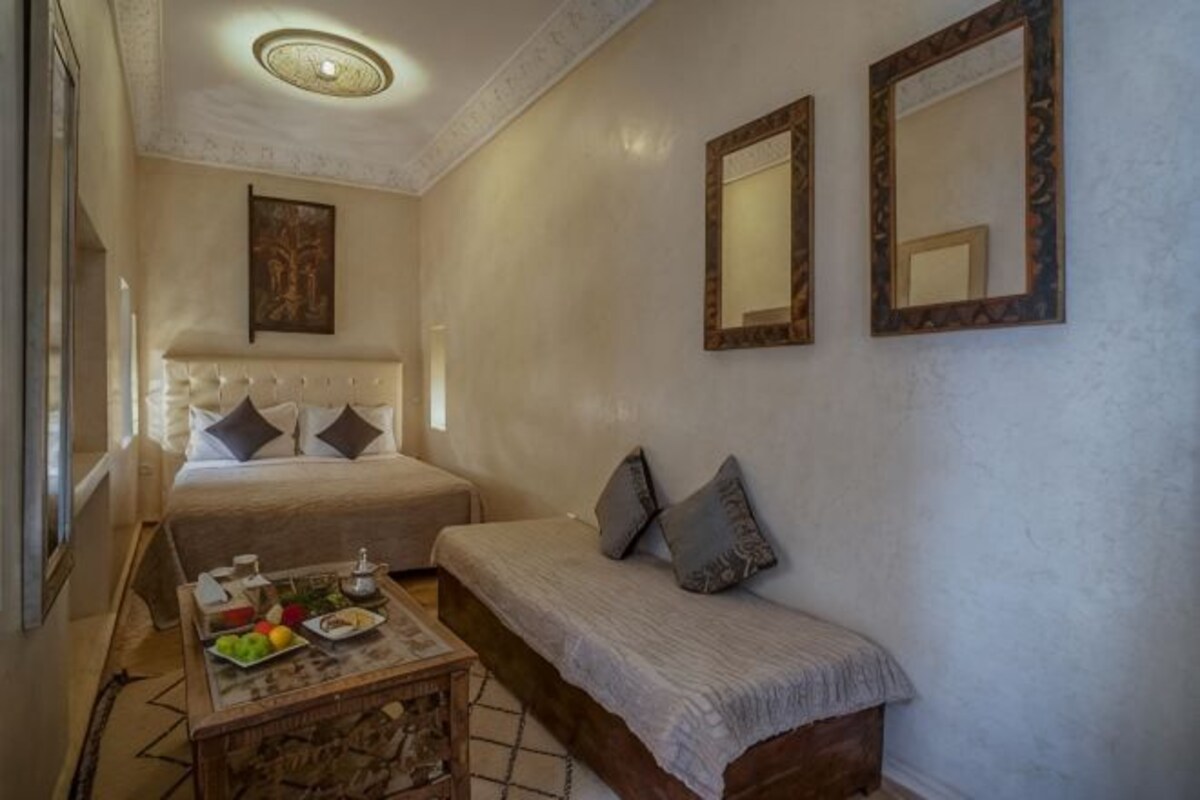 A bedroom with a bed, a sofa, a small table with fruits and a bottle, and two mirrors on the wall.