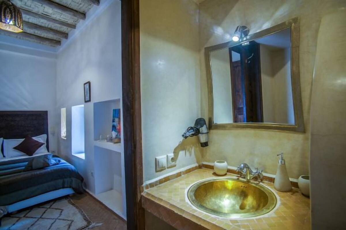 A bedroom with a bed, a bedside table, a nightstand, a window, and a bathroom with a sink and mirror.