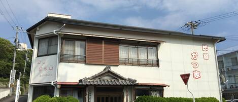 A two-story building with a traditional Japanese architectural style, featuring a tiled roof and a small front garden with two bushes.