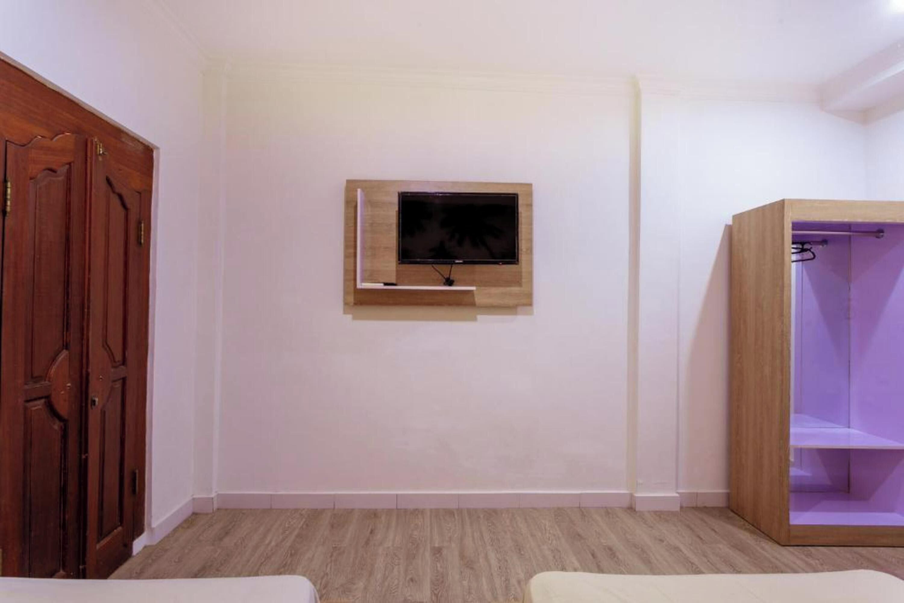 A room with a wooden wardrobe, a mounted television, and a wooden cabinet with shelves.
