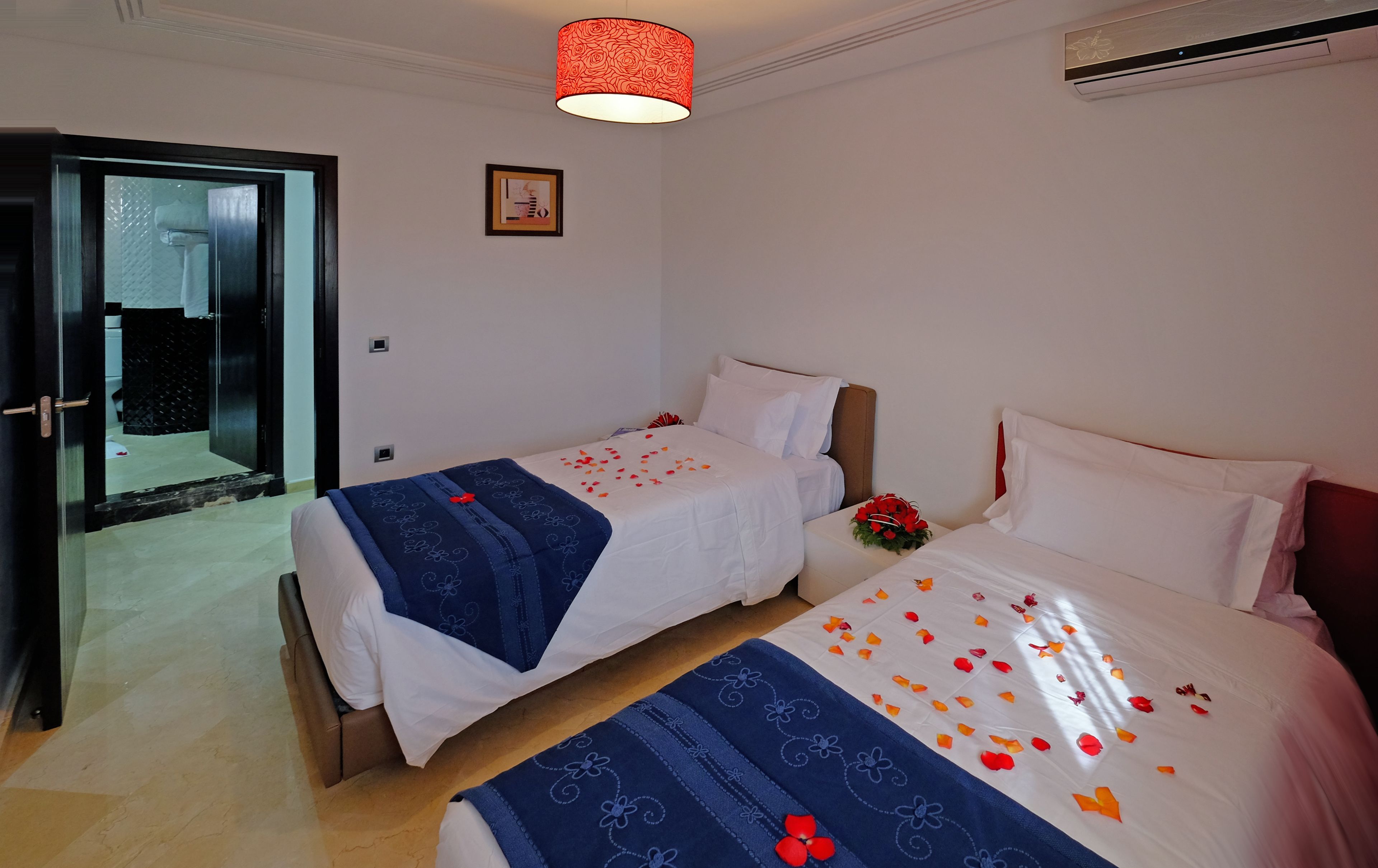 Habitación de hotel con dos camas, una puerta de cristal que da al baño y una lámpara roja con diseño estampado.