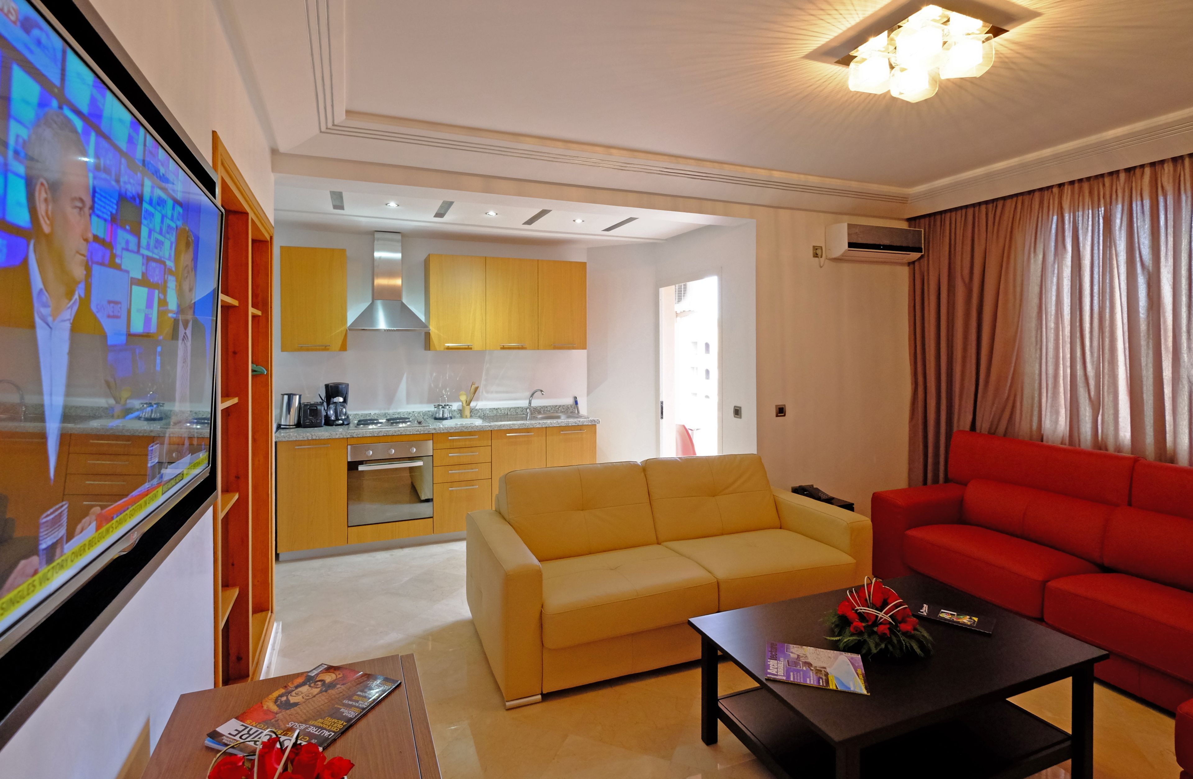 A living room with a red sofa, a yellow sofa, a coffee table with a magazine and flowers, and a TV displaying a news program.