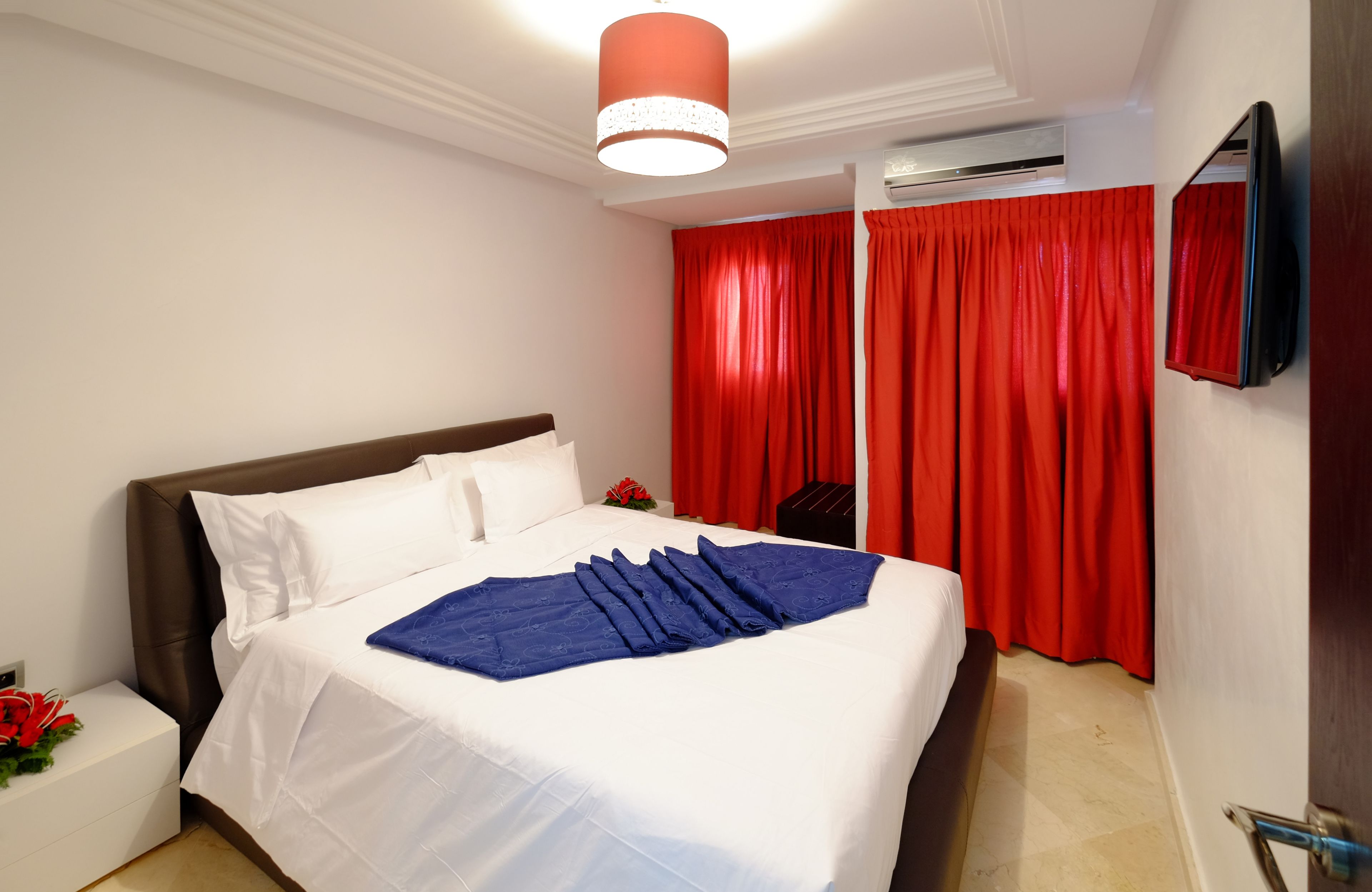 Uma cama bem arrumada com roupa de cama branca e azul, uma cortina vermelha e uma TV fixada na parede.