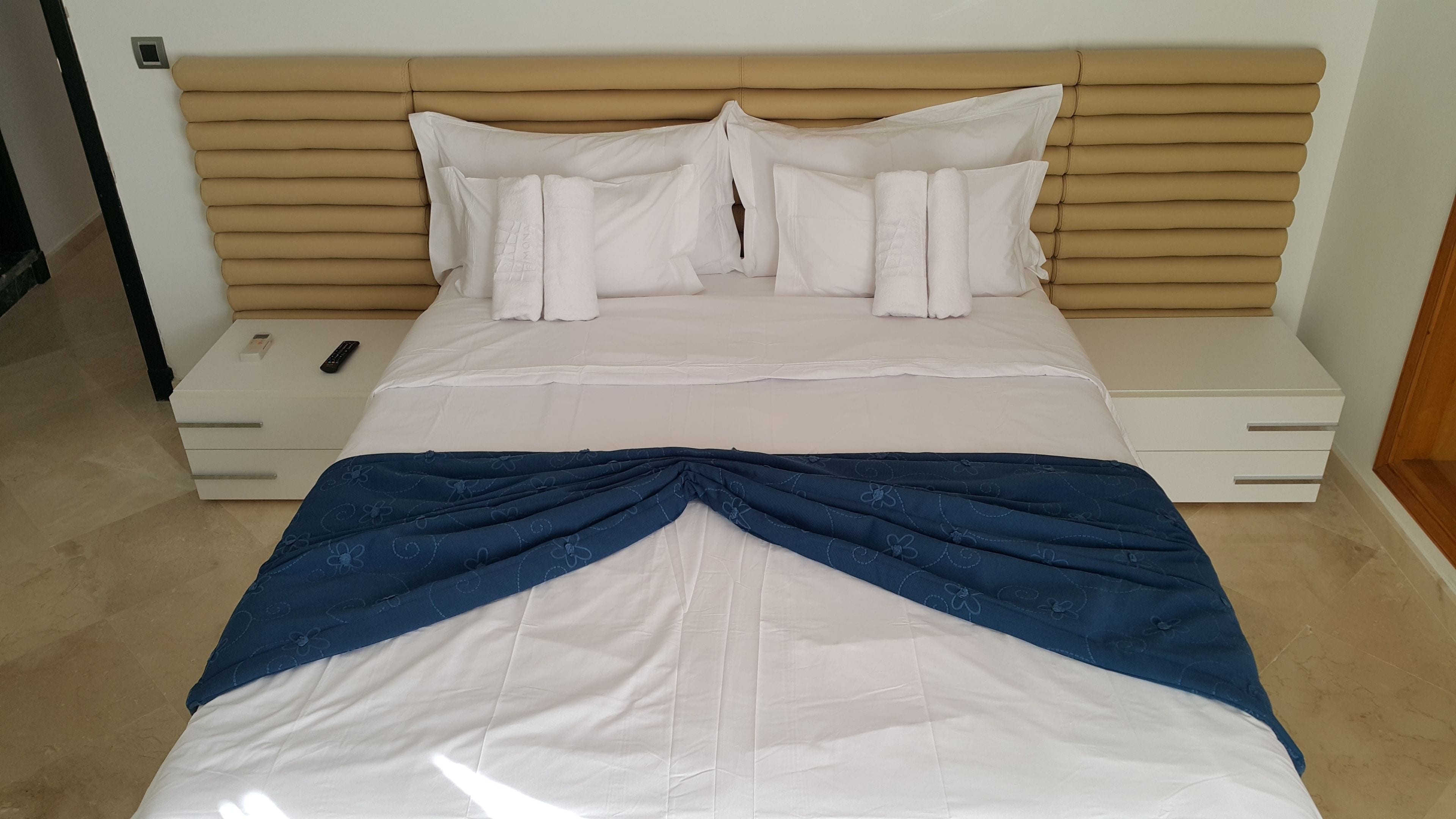 A neatly made bed with a white and blue blanket, a wooden headboard, and a bedside table with a remote control.