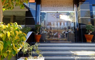 Una entrada moderna de edificio con puerta de cristal, plantas en macetas y un cartel con mensajes de bienvenida en varios idiomas.