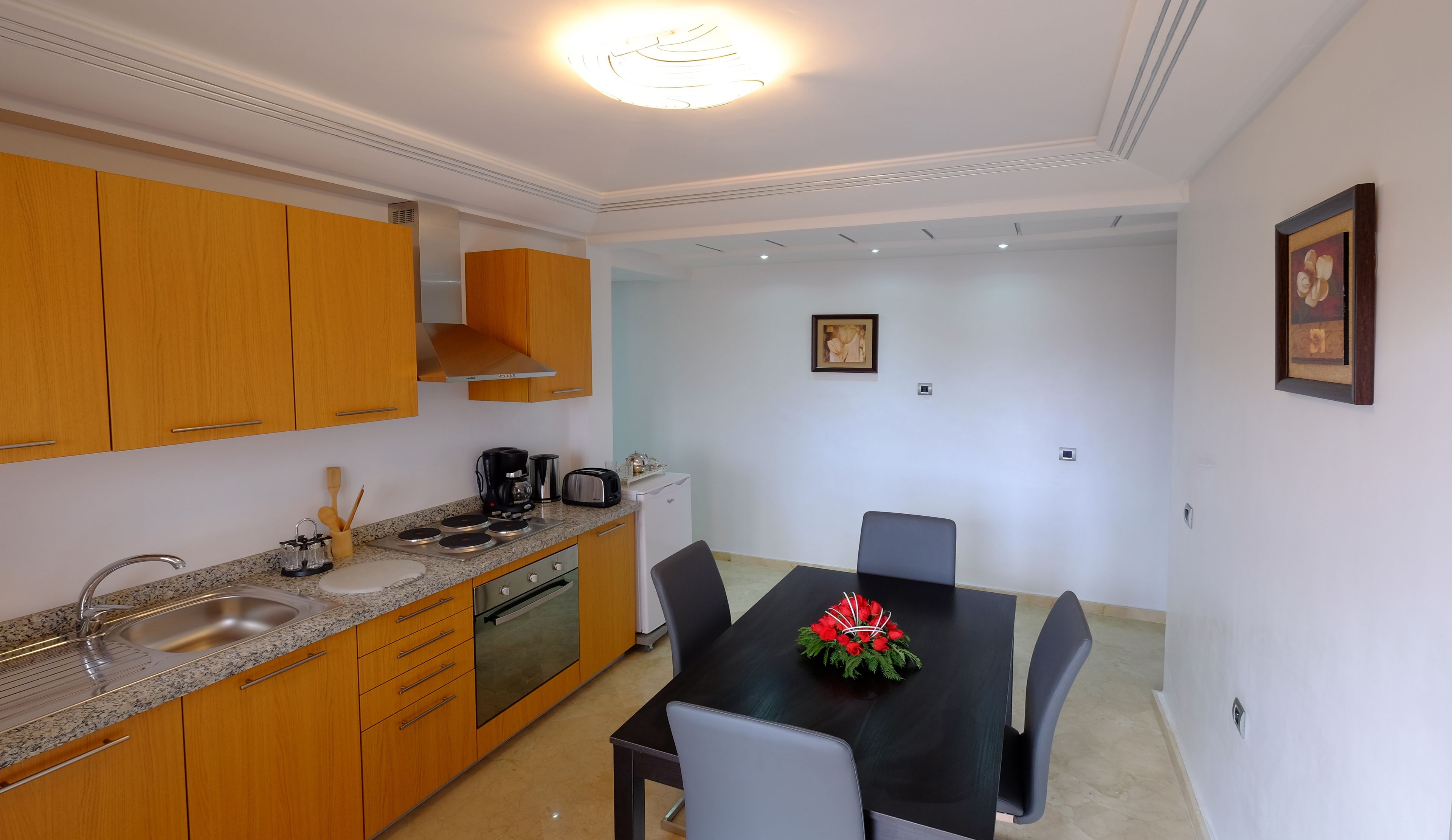 A modern kitchen with wooden cabinets, a granite countertop, and stainless steel appliances. There is a dining table with chairs and a vase of red flowers.
