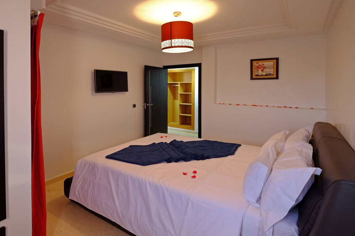 A neatly made bed with a white and blue bedspread, a red curtain, a red and white lamp, a framed picture, and a wooden wardrobe.
