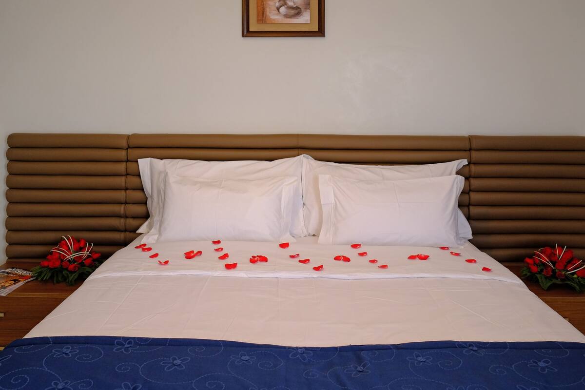 A neatly made bed with white linens, a brown headboard, and red rose petals scattered on the bed.
