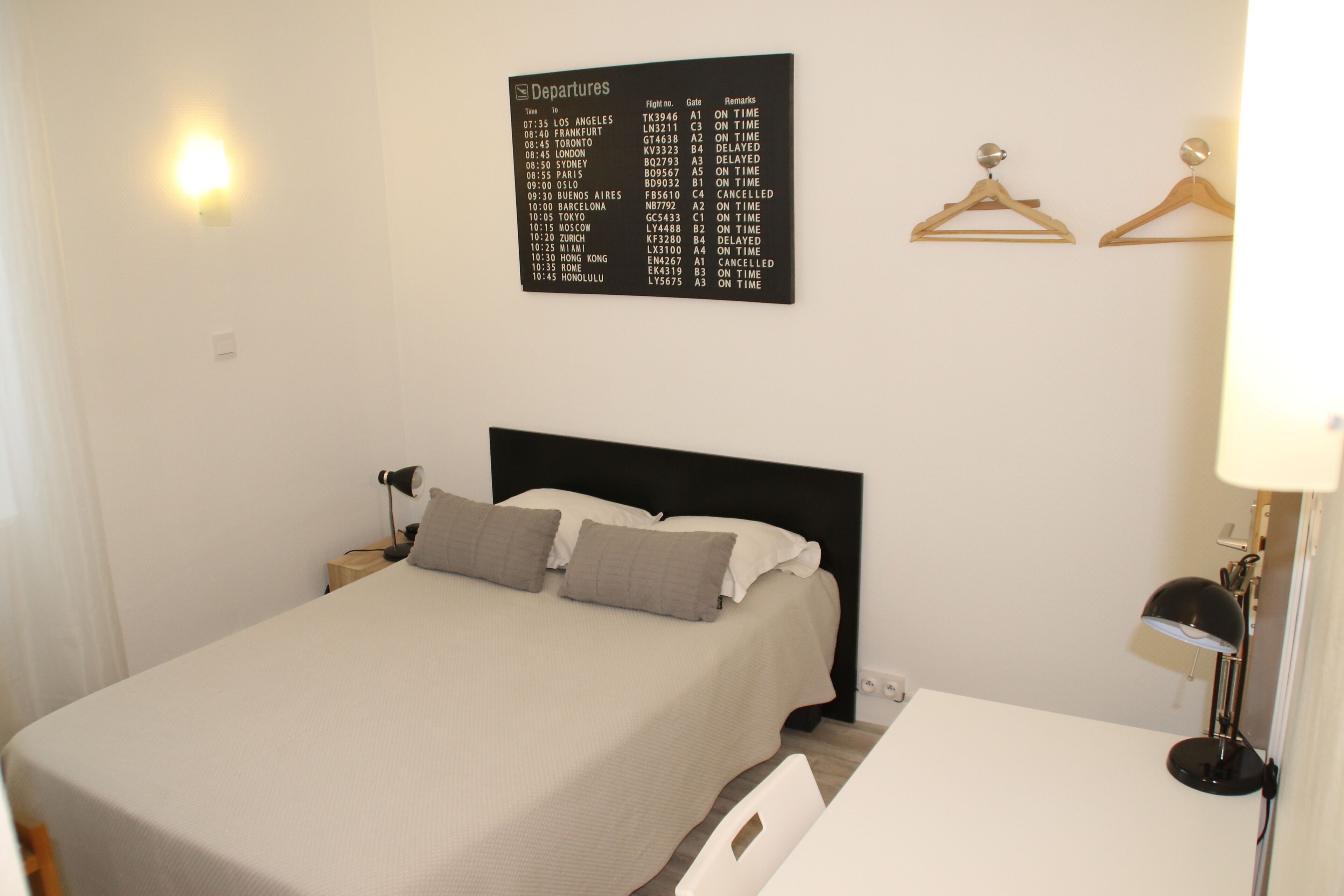 Una cama bien hecha con un cabecero negro, un panel informativo de salidas en la pared y una mesita de noche con una lámpara.