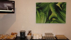 Una mesa con un bufé de panes, café y platos, una cafetera y un televisor mostrando un noticiero.