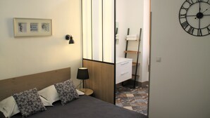 Un dormitorio con cama, almohadas, mesita de noche con lámpara, cómoda, espejo y un reloj de pared.