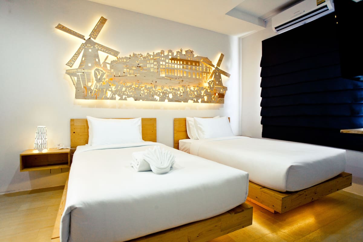 A hotel room with two beds, a wooden floor, a wall-mounted windmill sculpture, and a bedside table with a lamp.