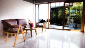 Una sala moderna con un sofá de cuero, sillas de madera, una mesita redonda y un amplio ventanal de vidrio que da a un área exterior con vegetación.