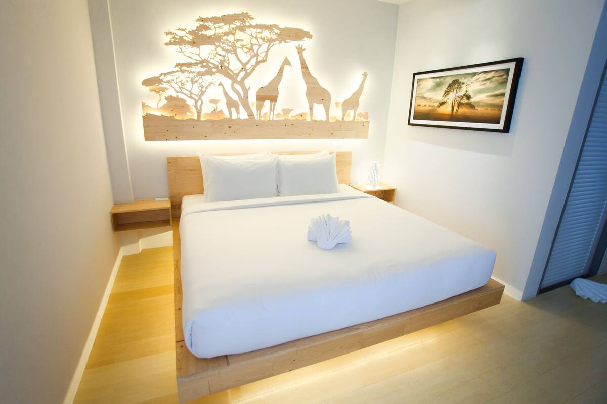 A neatly made bed with white linens and a folded towel, a wooden headboard, a wall-mounted artwork of giraffes, and a framed picture of a landscape.
