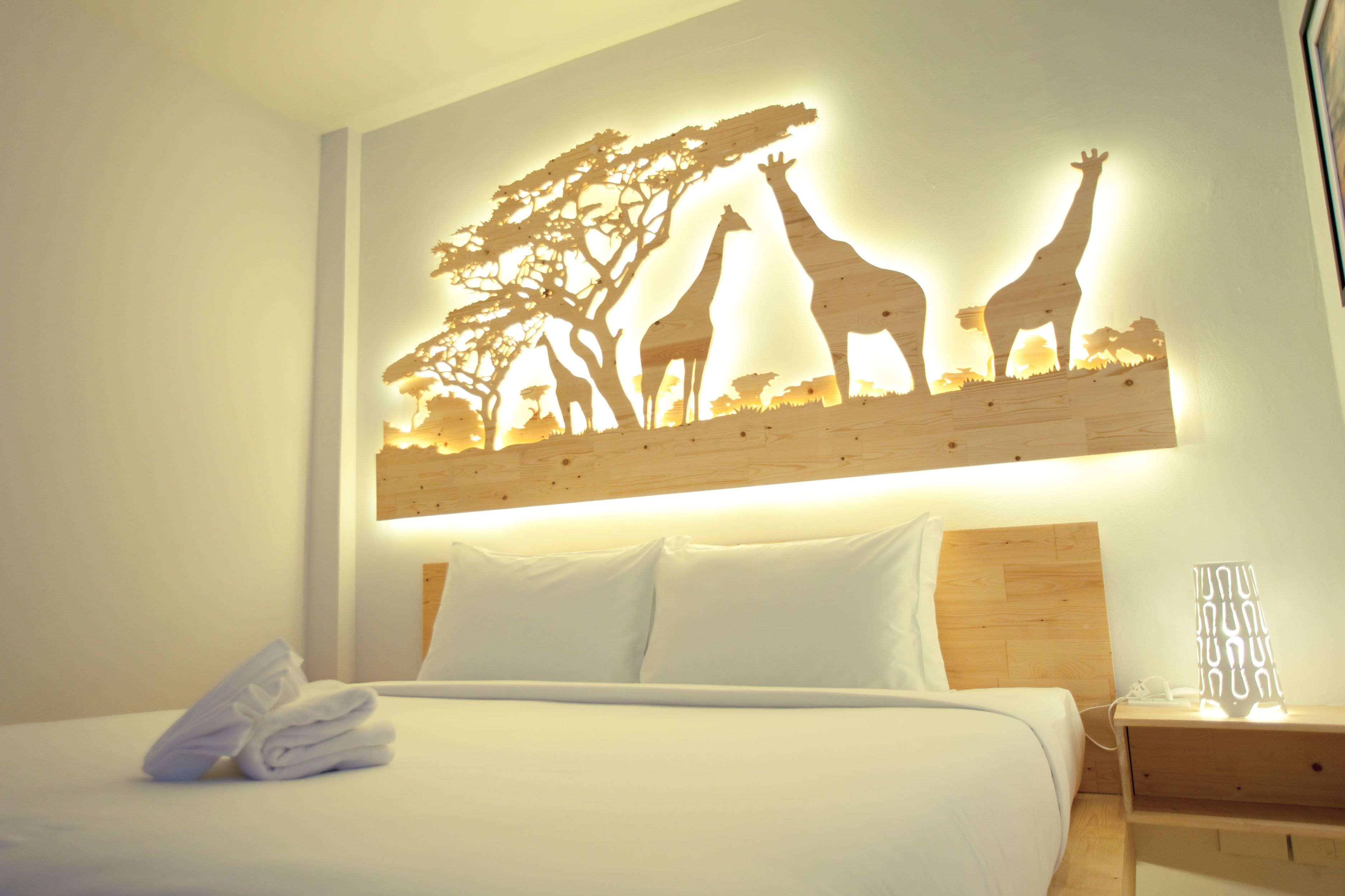 Habitación de hotel con cama, almohadas, una toalla doblada con cuidado, una mesita de noche con lámpara y una obra de arte con jirafas y árboles.