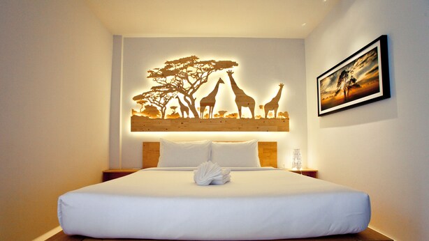 Una cama bien tendida con sábanas blancas, cabecera de madera y una obra de arte en la pared que representa jirafas y árboles.