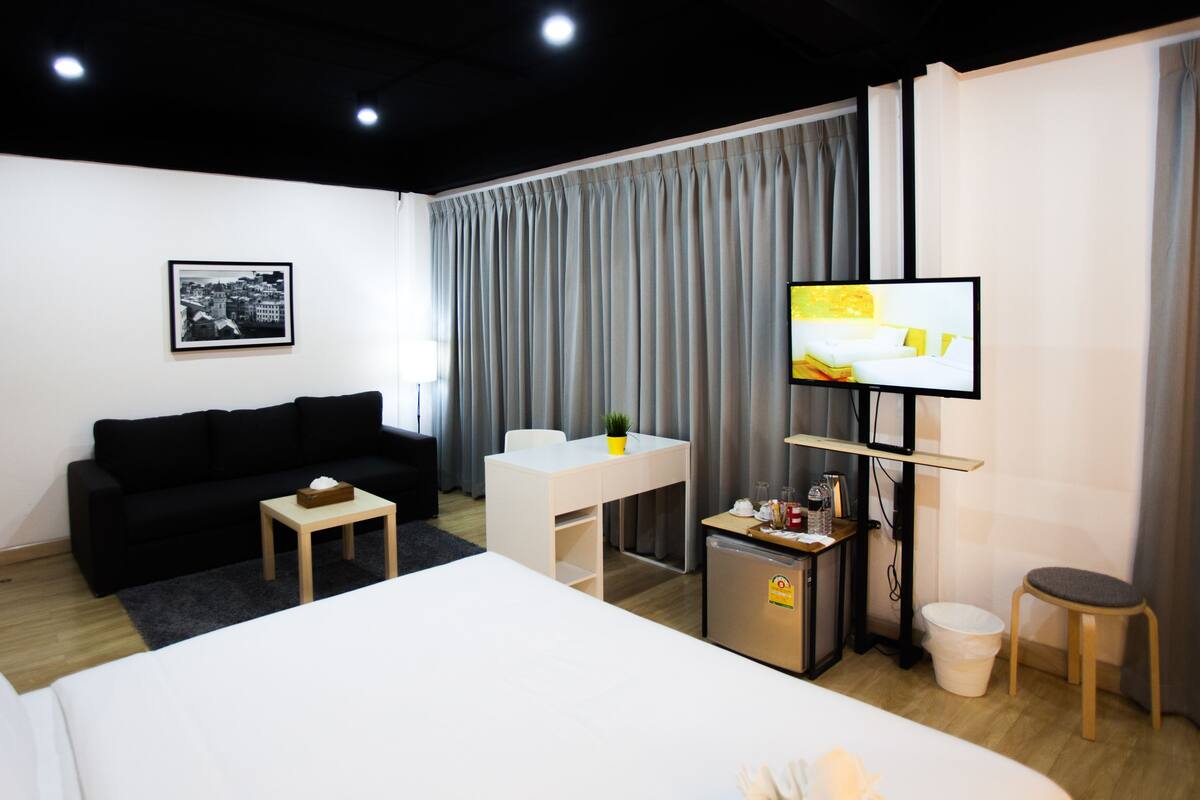 A modern hotel room with a bed, a black sofa, a small table, a TV, and a window with curtains.