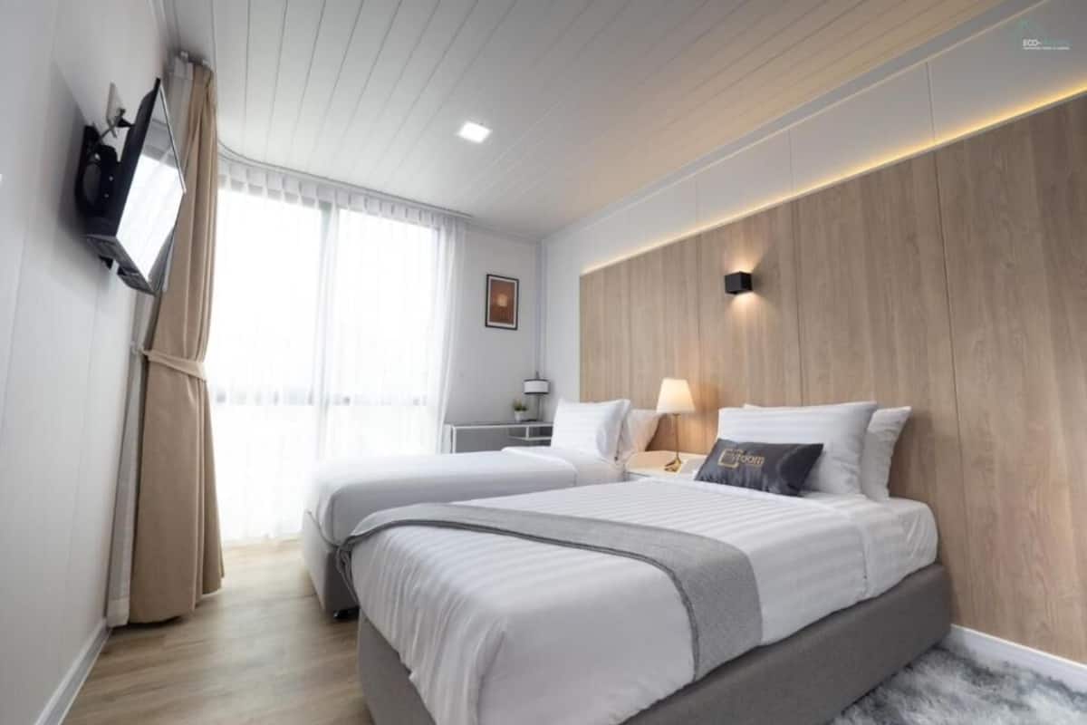 A modern hotel room with two beds, a flat-screen TV mounted on the wall, a wooden accent wall, and a large window with curtains.