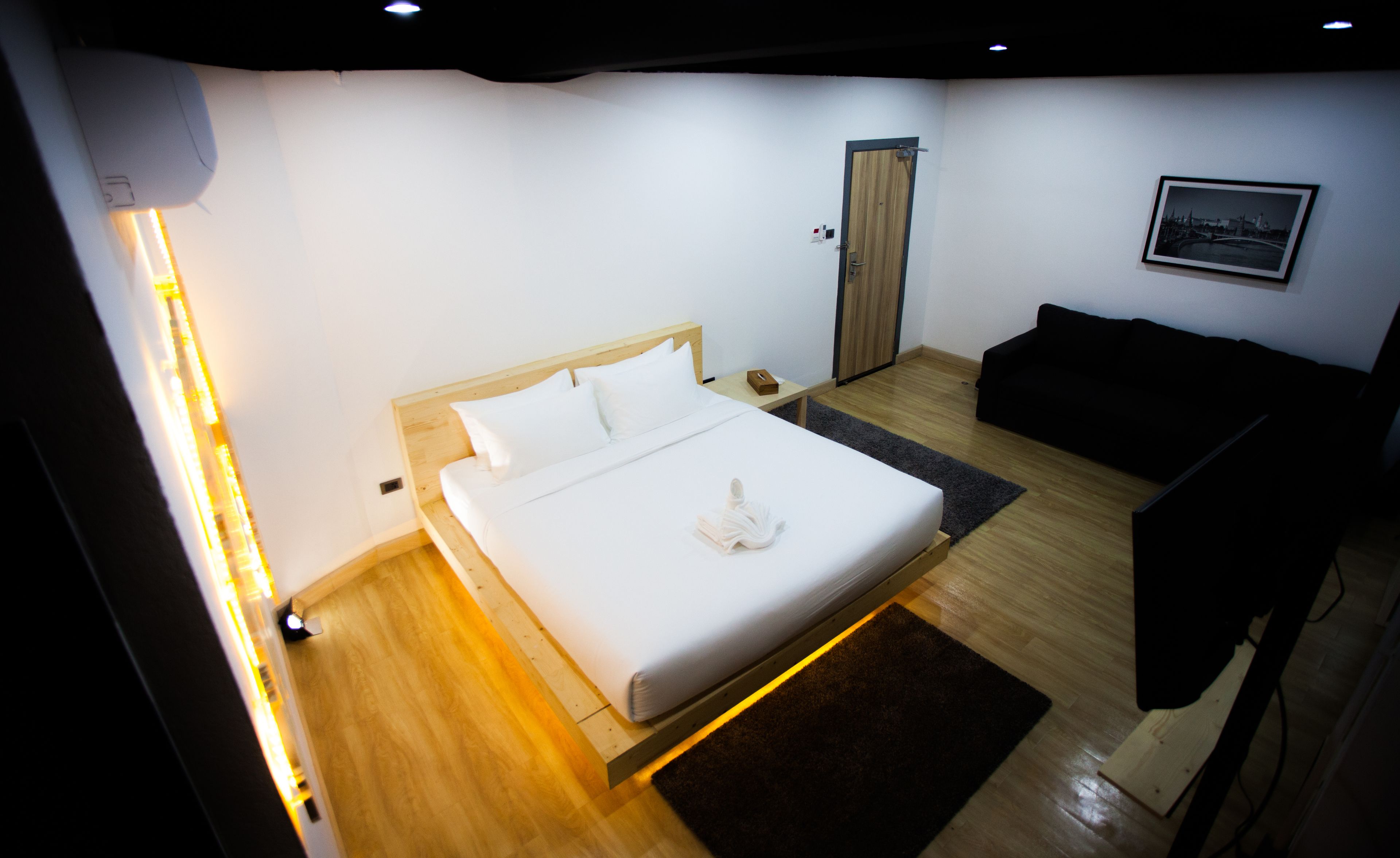 A modern hotel room with a wooden bed, a black sofa, a framed picture on the wall, and a wooden floor.