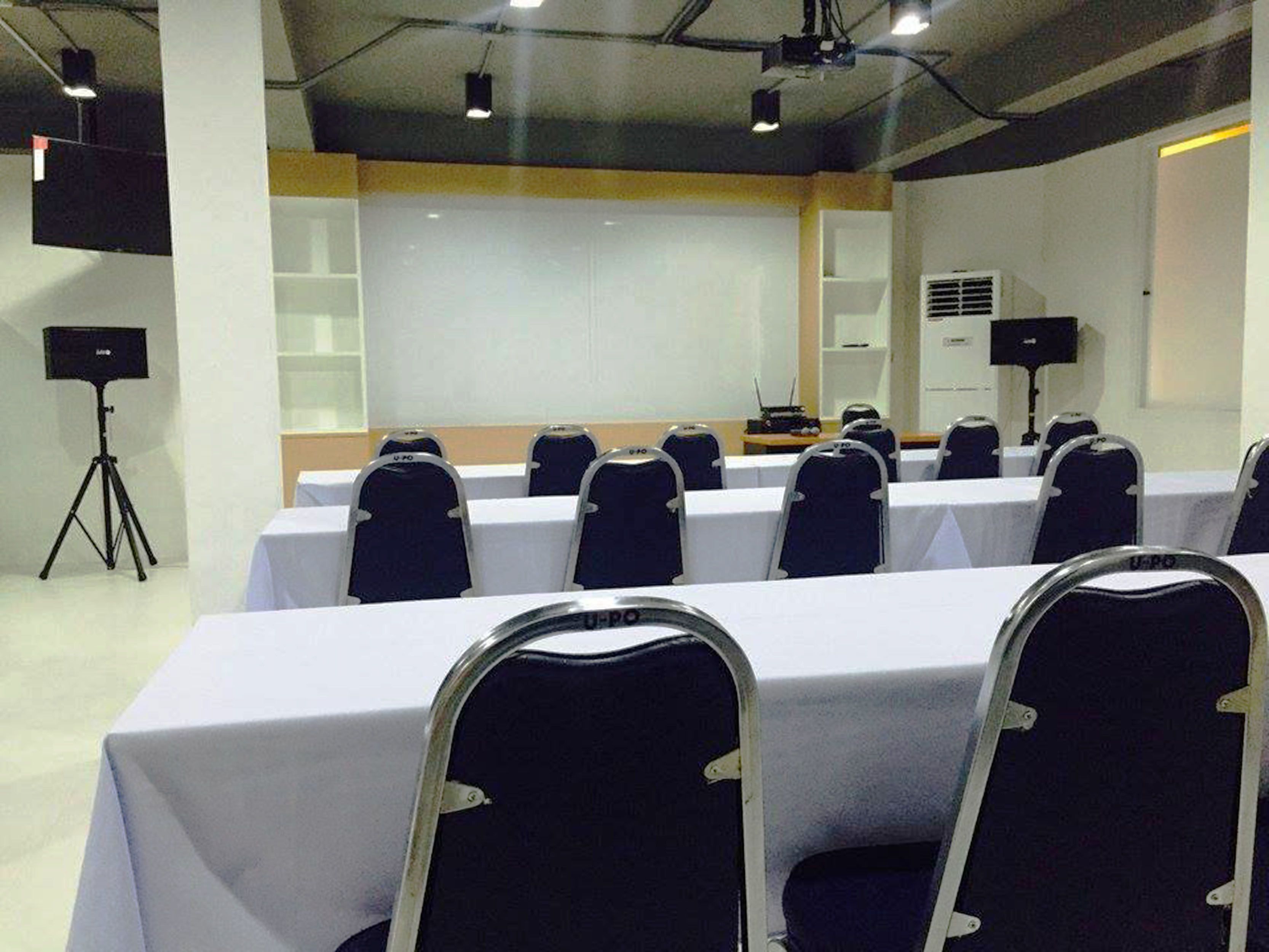 Une salle de conférence avec des rangées de tables et de chaises, un projecteur et un tableau blanc.
